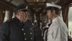 Im Speisewagen eines historischen Zuges. Links steht ein älterer, korpulenter Mann. Er trägt eine blaue Uniform mit doppelter Knopfreihe und eine Offiziersmütze. Rechts steht ein junger Mann in einer weißen Uniform mit weißer Offiziersmütze. Der ältere Mann schimpft mit dem jüngeren Mann. Der jüngere Mann schaut verlegen weg.