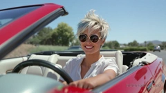 A young woman sits in the driver&rsquo;s seat of a red convertible. She has a short, tousled pixie cut with blue streaks, and is wearing sunglasses with a golden rims. She smiles as she rests her left arm on the car door.