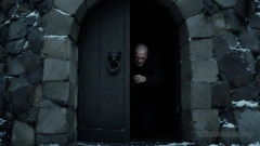 The image depicts a dark, eerie Winter scene featuring the stone structure of a castle with a large, arched wooden door. The door is slightly ajar, revealing a dimly lit older man with a serious expression. The stone wall surrounding the door is composed of large, irregular stones, some of which are dusted with snow, adding to the cold and foreboding atmosphere. The door has a distinctive black metal ring handle shaped like an animal&rsquo;s head.