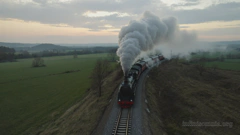 In der Dämmerung fährt ein Zug durch eine ländliche Landschaft. Ganz vorne befindet sich eine Dampflokomotive, gefolgt von mehreren Waggons, von denen einige in Rauch gehüllt sind. Du stehst leicht erhöht und blickst direkt auf den Zug.