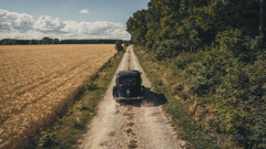 Ein schwarzer Oldtimer fährt auf einer Landstraße zwischen einem Feld und einem Wald. Das Wetter ist sommerlich und angenehm, aber in der Ferne ziehen bereits Gewitterwolken auf.