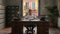 A young man is sitting behind a desk in an office, with filing cabinets against the wall and a plant in the corner. Everything looks a bit worn. From the window behind him, there is a terracotta-coloured wall with balconies.
