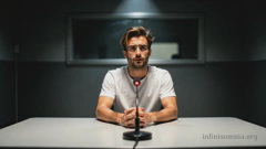 Dian in einem dunklen Verhörraum. Er sitzt im Scheinwerferlicht und wirkt besorgt.  Vor ihm, auf dem Tisch, steht ein Mikrofon mit einem roten Licht, bereit zur Aufnahme. Hinter ihm befindet sich ein Einwegspiegel.