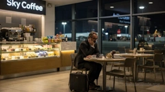 Late at night, a man is waiting in an empty airport café. He looks tired and frustrated. There are several coffee cups and empty plates on the table. A briefcase is sitting next to him.