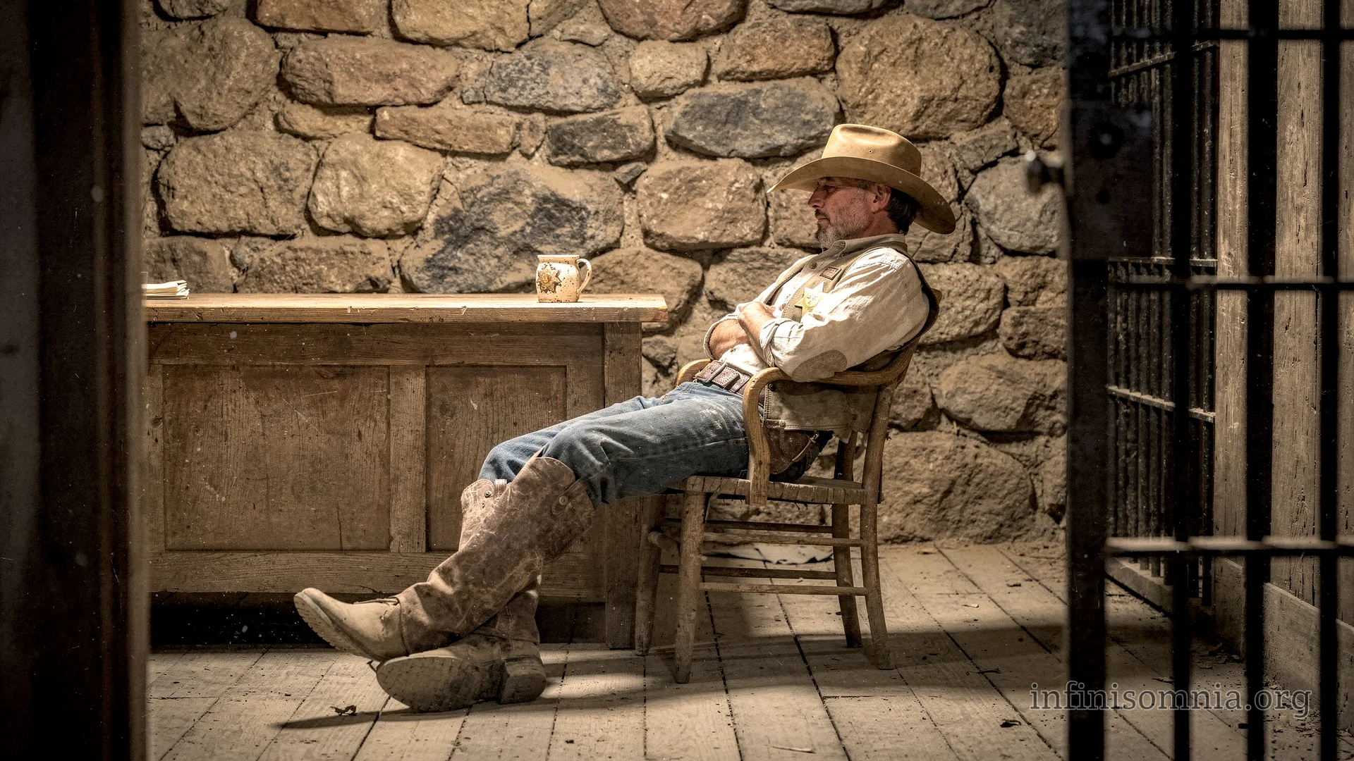A sheriff&rsquo;s office. Stone walls, a desk with a mug, and to the side, the barred door of a prison cell. The sheriff is sitting in a chair with his legs stretched out, dozing off.
