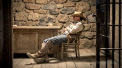 A sheriff&rsquo;s office. Stone walls, a desk with a mug, and to the side, the barred door of a prison cell. The sheriff is sitting in a chair with his legs stretched out, dozing off.
