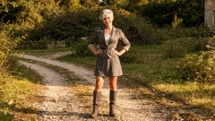 A gravel path leads through a neglected park. Miray is standing on the path wearing a grey dress and boots. She has her hands on her hips and is smiling at the camera. A wall and the edge of a building are visible behind her.