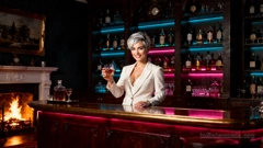 An old-fashioned room with a crackling fire in the hearth. A bar counter stands in the room, looking somewhat out of place. On the wall is a shelf of spirits, illuminated by blue and pink strip lights. Miray is behind the bar. She is wearing a bartender&rsquo;s white suit, holding a whisky glass and smiling. (AI generated image)