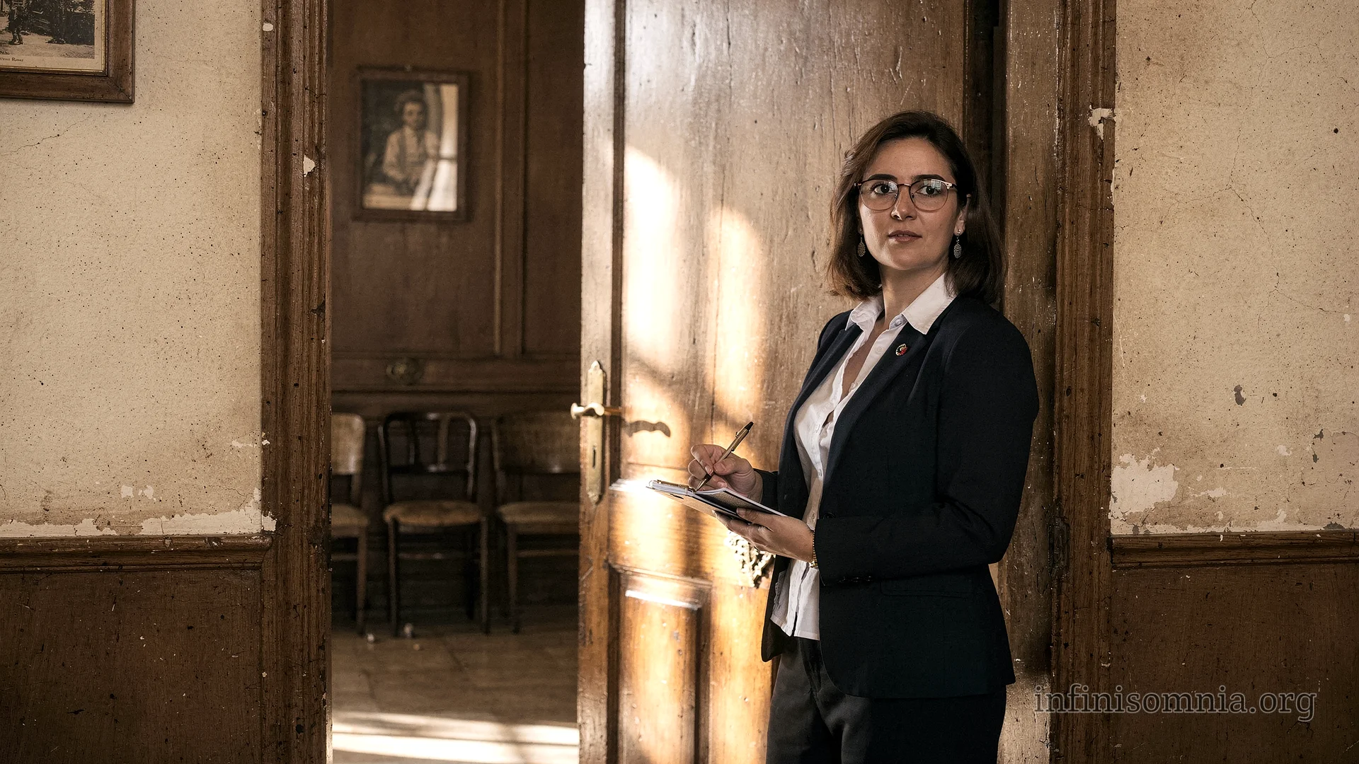A somewhat run-down office. Standing in the doorway to the waiting room is the secretary, Clara Montserrat. She has a notebook and a pen in her hand.
