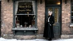 A brick building front with a door and a small shop window. On the window, it says &ldquo;Miray & Co. Inquiry Office&rdquo;. Miray is standing at the door. She is wearing a long black coat with a high collar. It is winter, and the street is covered in snow. (AI generated image)