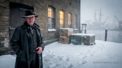 A man in a black coat and hat is standing in front of an old warehouse. He seems to be waiting for someone. The warehouse is situated on a quay. In the background, a crane and a sailing ship are visible. It is cold and misty. There is snow on the ground. (AI generated image)