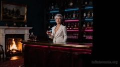Ein altmodisches Kaminzimmer mit einem prasselnden Kaminfeuer. Im Raum steht – etwas deplatziert – ein Bartresen. An der Wand ist ein Regal mit Spirituosen, beleuchtet mit blauen und pinken Leuchtstreifen. Hinter dem Tresen steht Miray. Sie trägt den weißen Anzug einer Barkeeperin. Sie lächelt und reicht ein Whiskyglas an, mit den Worten: „Guten Abend, Doktor Hilton.“ (KI-generiertes Bild)